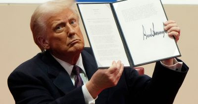 Donald Trump holding up a signed executive order document at a press briefing.